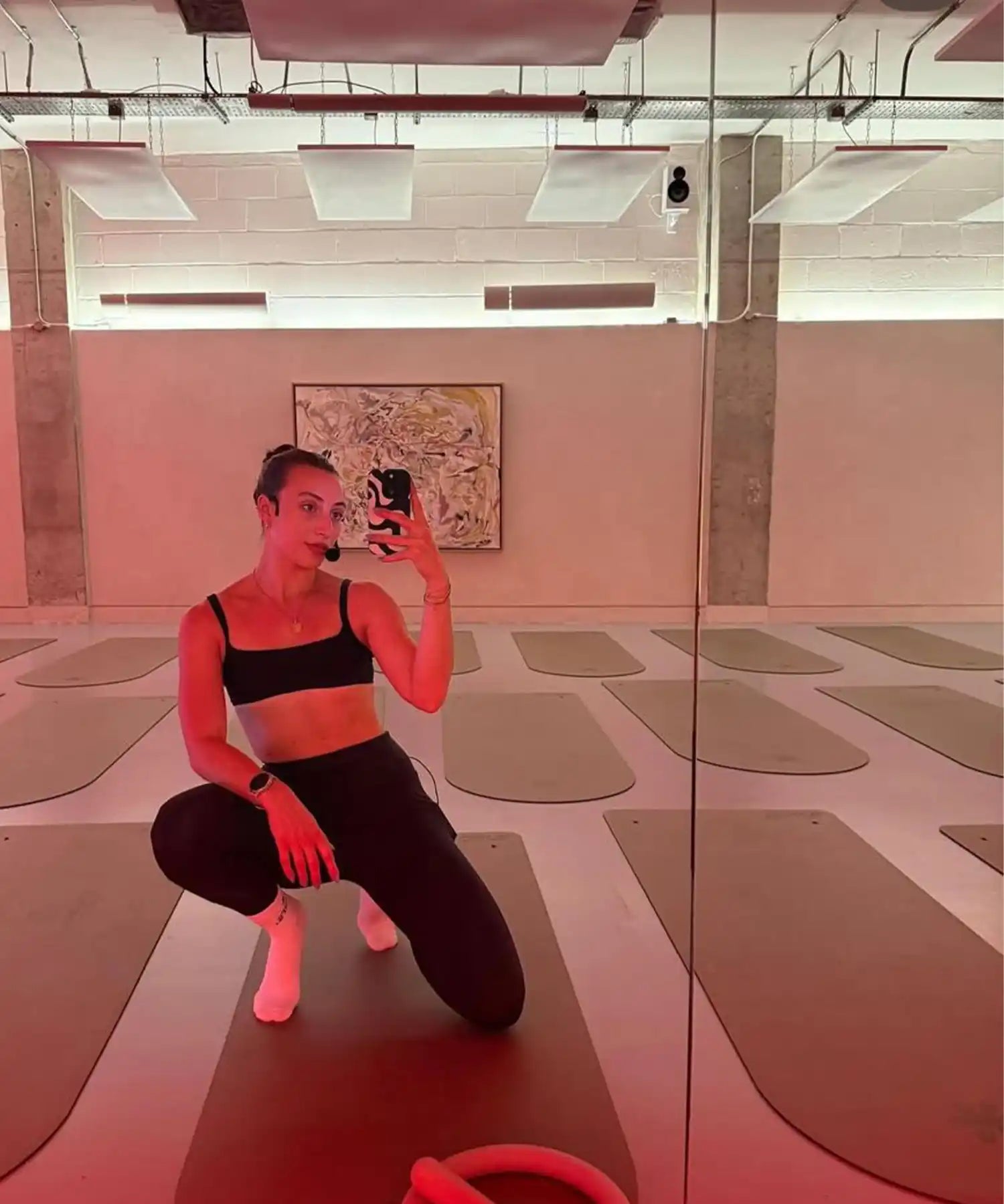 Mirror view of a participant kneeling on a mat inside YOURS Studio London, infrared heating panels overhead, red ambient glow, studio interior visible.