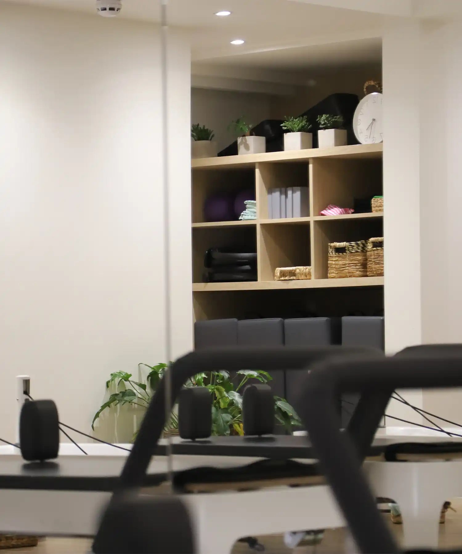 Storage area inside Reformer Revolution studio with neatly arranged Pilates props, yoga accessories, rolled towels, plants, baskets and a wall clock, seen through the reformer machines in the foreground.