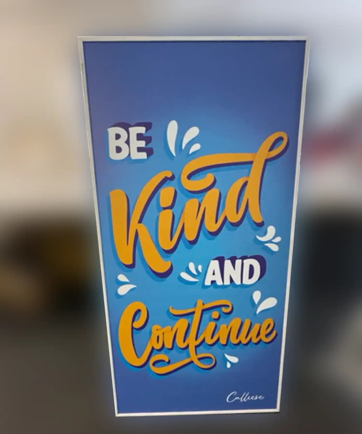 A custom infrared heating panel with a bold and uplifting message in hand-lettered typography. Against a gradient blue background, the text reads: “Be Kind and Continue” in bright orange, white, and purple lettering, with decorative droplets and a signature that reads “Calluse.”