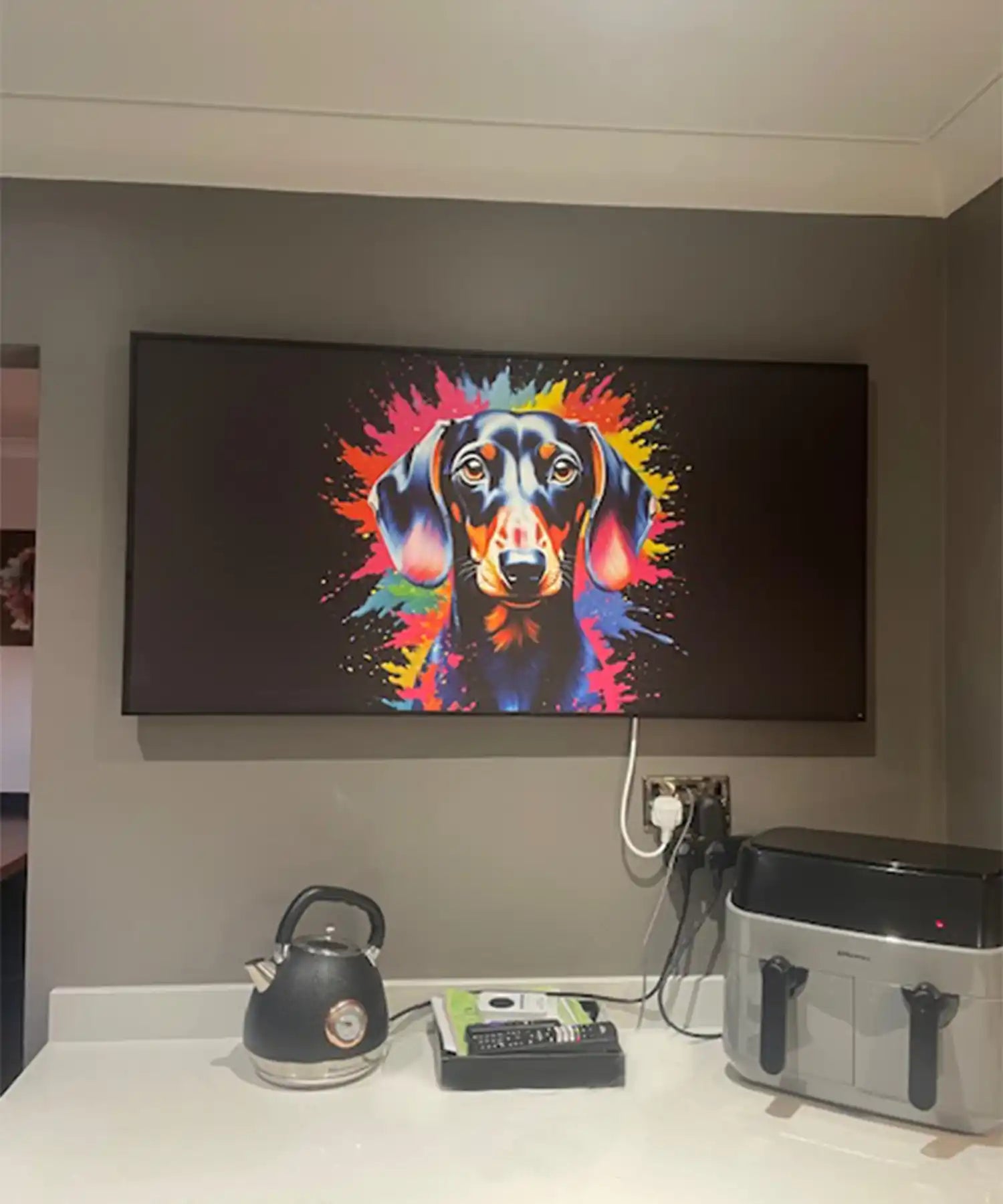A custom infrared heating panel mounted in a modern kitchen, featuring a bold, colourful digital portrait of a dachshund with splashes of neon pink, orange, blue, and yellow exploding from behind. Below, a kettle, phone, and air fryer sit on the counter.