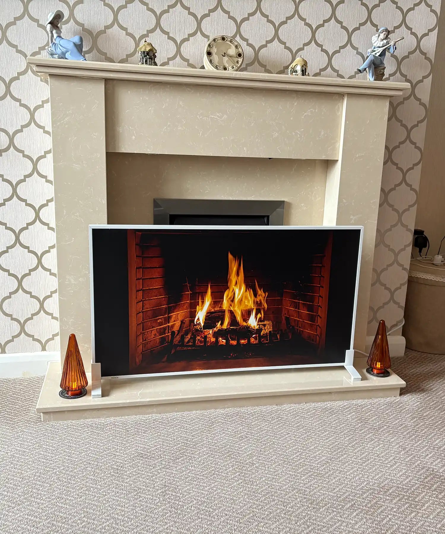 A realistic custom infrared heating panel depicting a roaring fireplace, set in front of a traditional cream-colored mantel. The panel emits the illusion of flickering flames, surrounded by two vintage amber glass lamps. The setup blends traditional décor with modern heating tech.