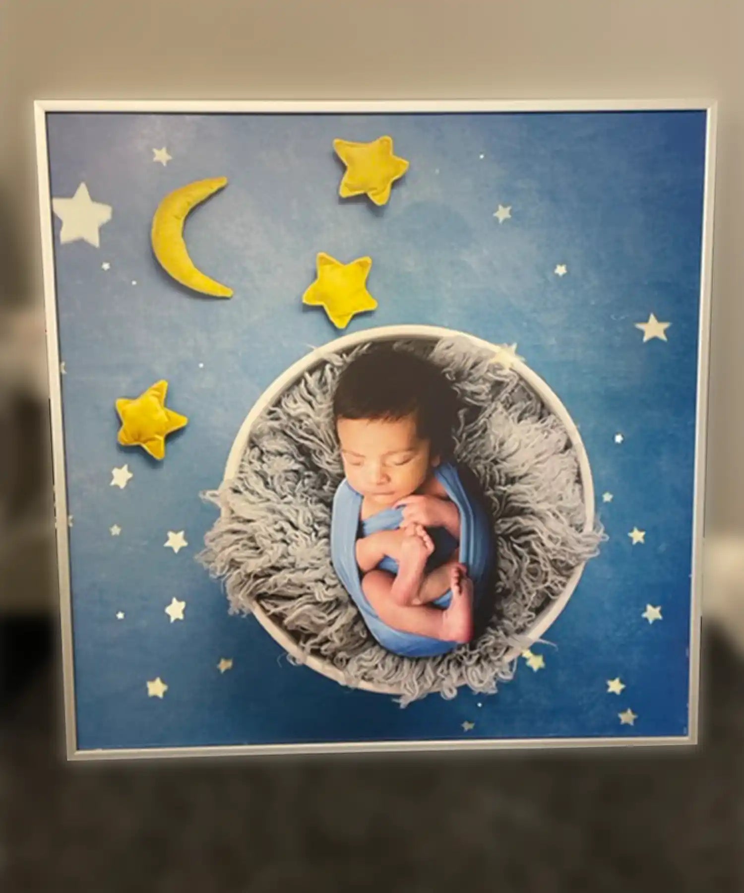 A custom infrared heating panel with a dreamy image of a newborn baby swaddled in blue, peacefully sleeping in a fluffy grey nest. The background is a deep blue night sky with scattered stars and plush yellow felt moon and stars surrounding the baby, giving a whimsical celestial vibe.