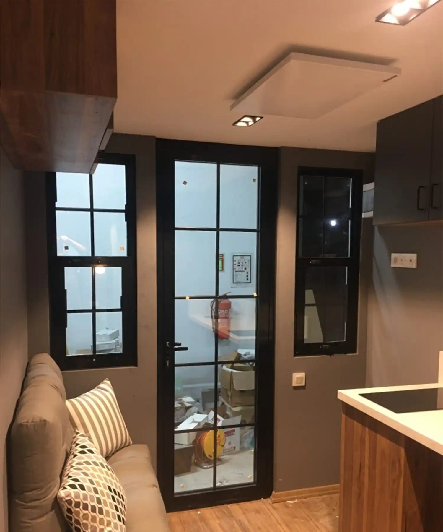 Kore Prime infrared ceiling heater in a compact, modern room with dark-framed windows and a grey sofa.