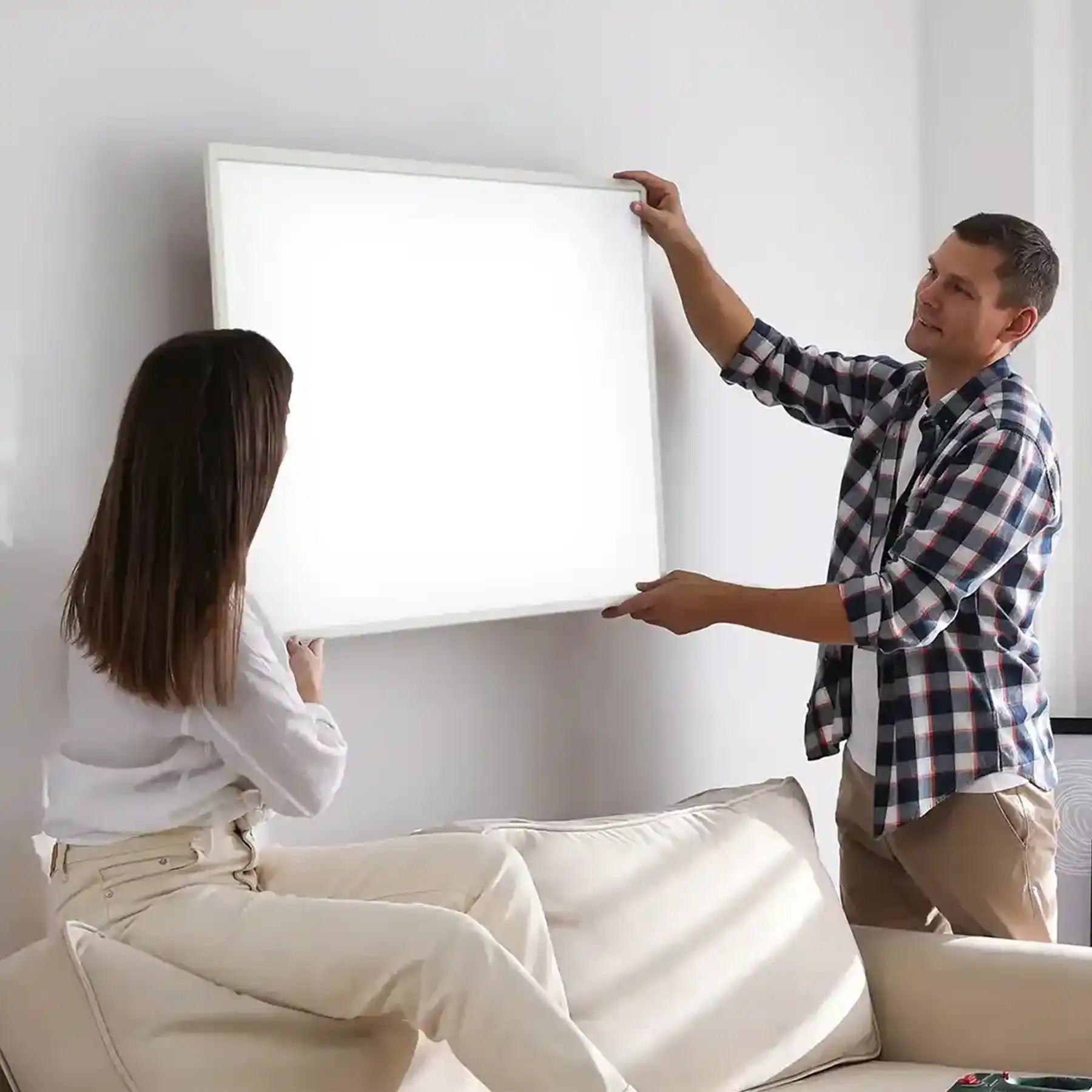 Couple mounting a Kiasa Classic infrared panel heater on the wall above a cream-colored sofa in a bright living room.