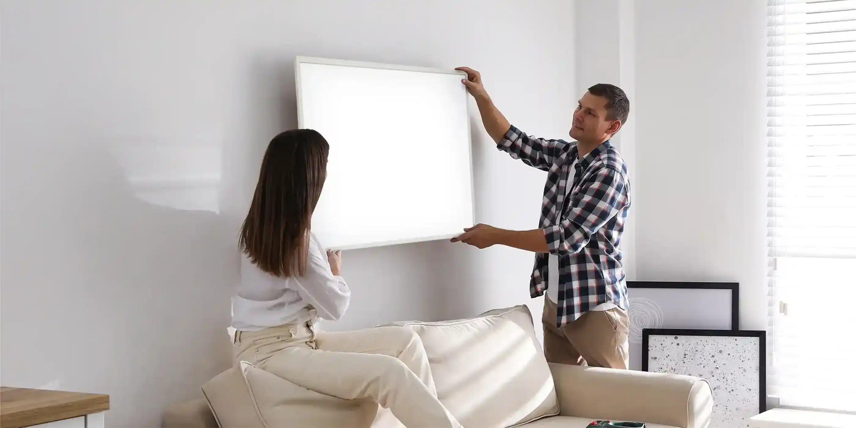 Couple mounting a Kiasa Classic infrared panel heater on the wall above a cream-colored sofa in a bright living room.