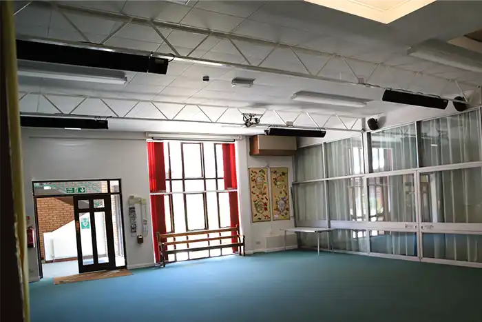 Large multi-purpose hall with high ceilings featuring multiple black infrared heater bars mounted overhead, showing spacious layout with large windows, carpeted floor and suspended ceiling framework in church with Kiasa IKON Heater Bars.