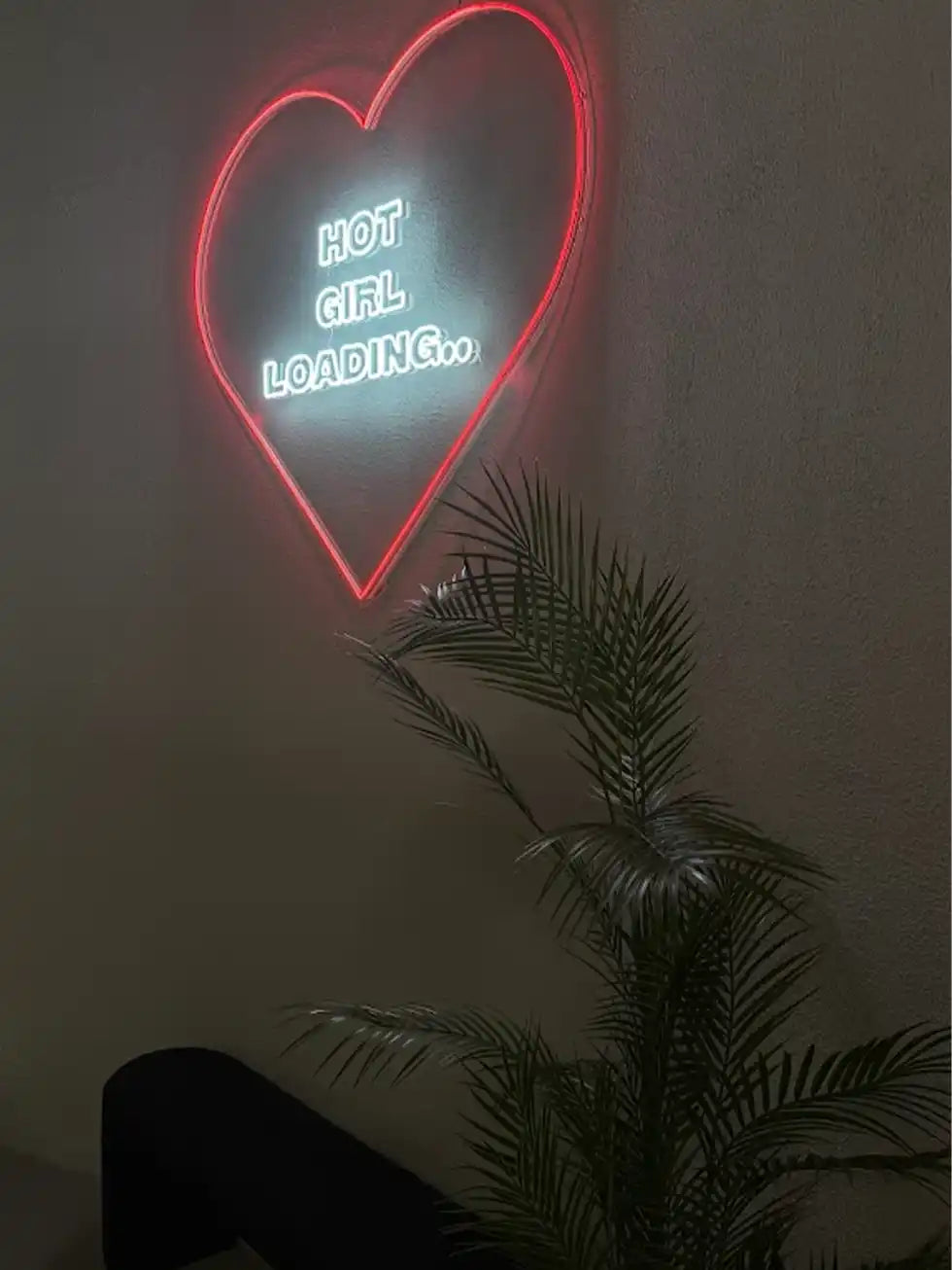 Neon sign with heart shape and text 'HOT GIRL LOADING...' on a dark wall with a plant in the foreground.
