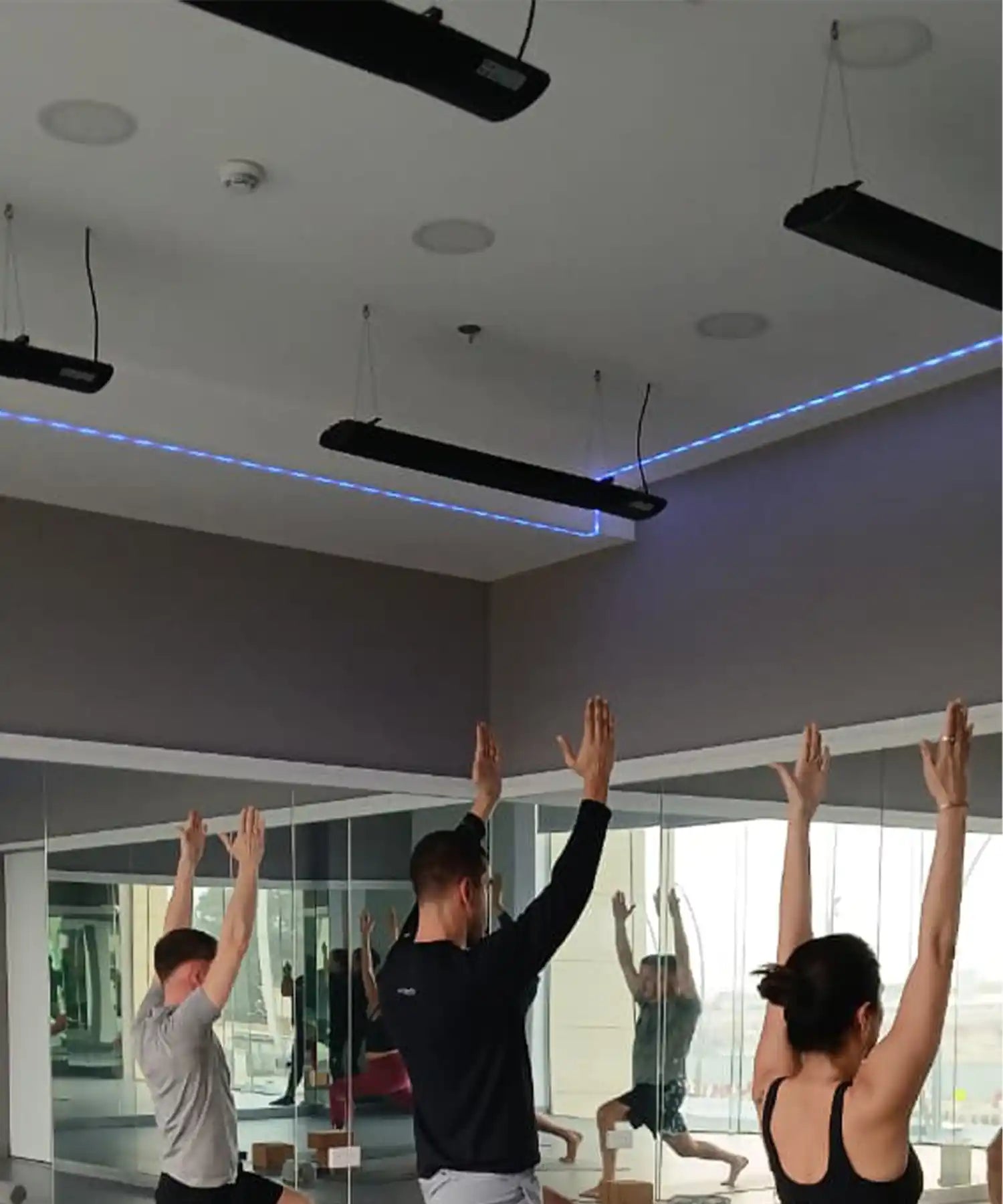 Fitness class performing standing poses in a mirrored studio with suspended infrared heater bars delivering consistent radiant heat across the space.