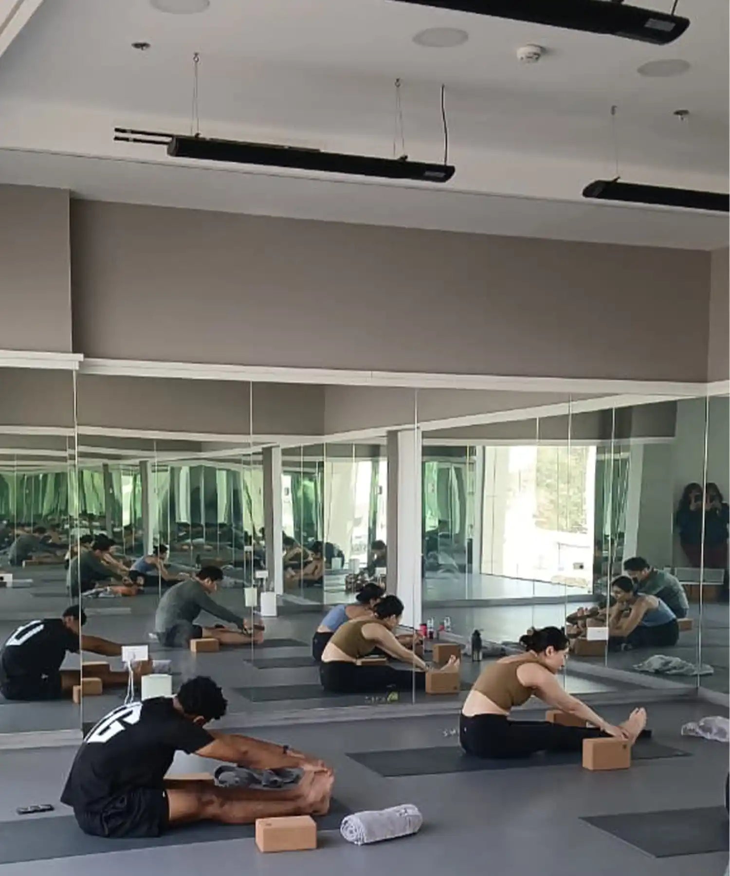 Group stretching class in a mirrored fitness studio with ceiling-mounted infrared heater bars delivering even, draft-free warmth during flexibility training.