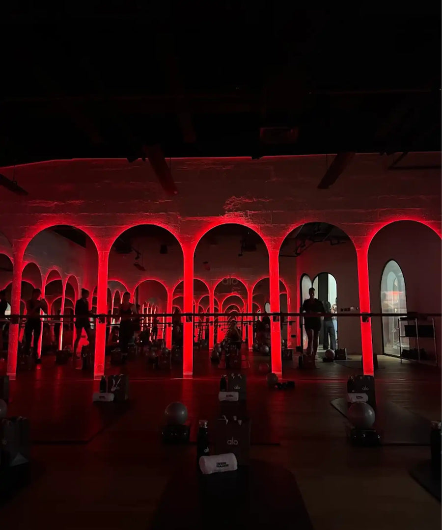 Women doing barre class in a hot-room studio at BRRN BARRE Dubai, with visible IR heater bars mounted overhead and warm ambient lighting.
