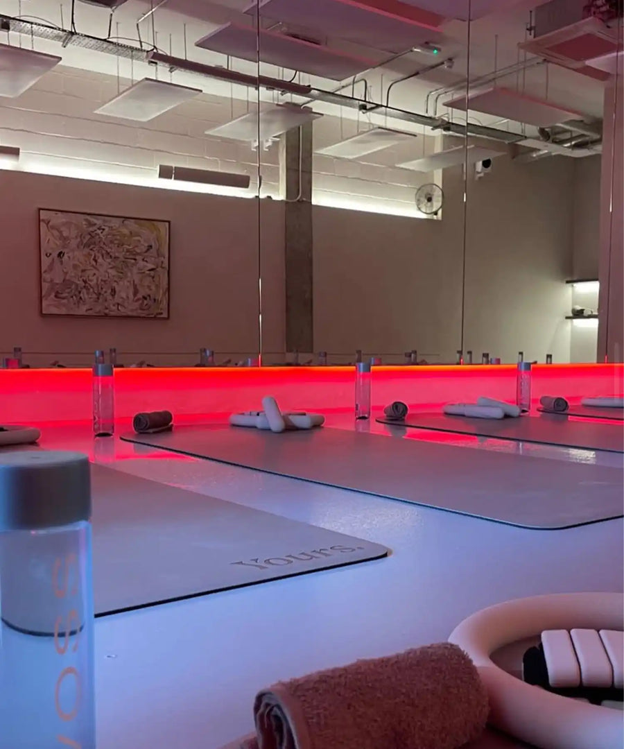Interior shot of YOURS Studio London showing empty yoga/Pilates mats arranged in room, ceiling-mounted infrared heating panels visible above, red-lit accent around mirror wall.