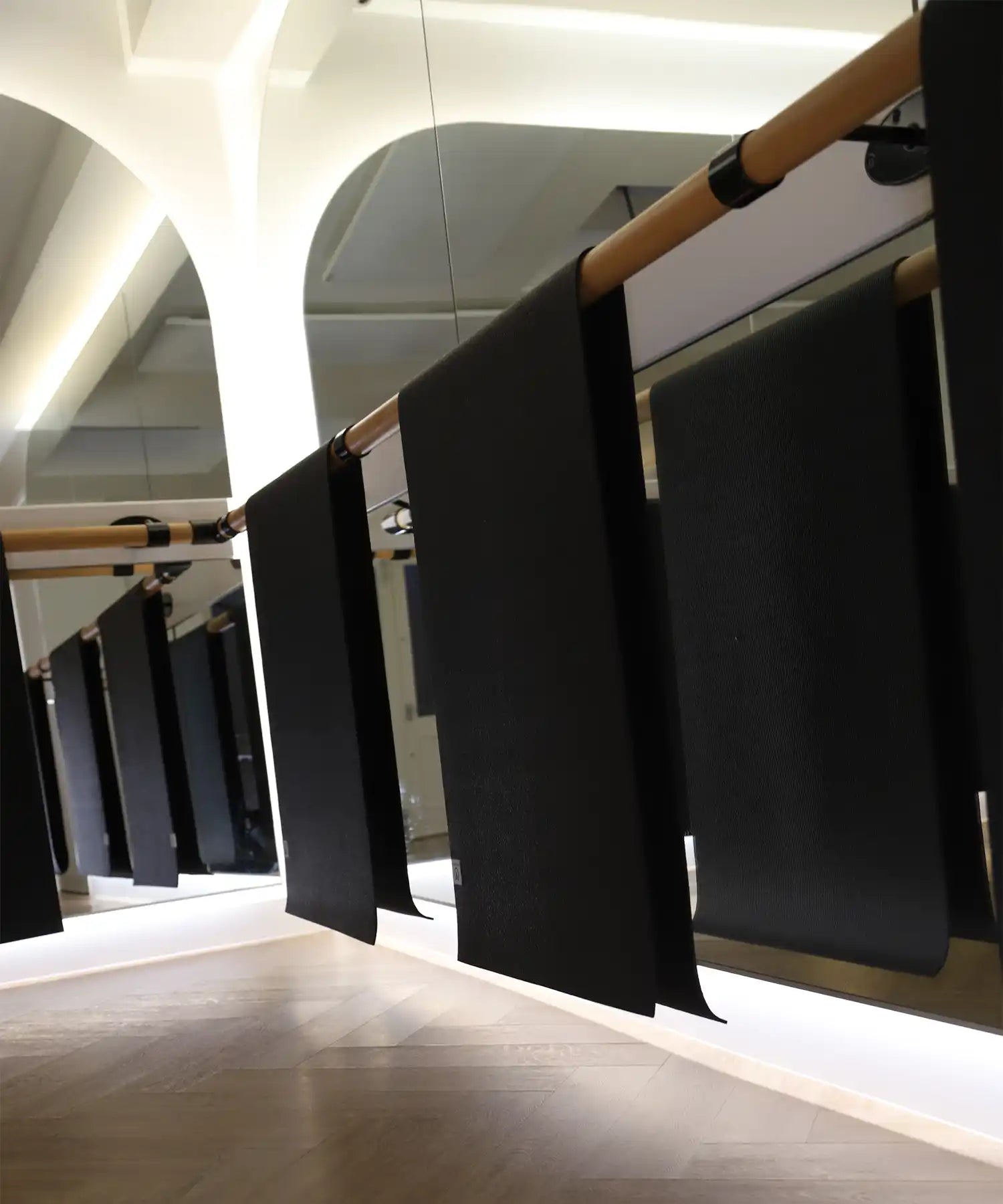 Close-up view of the barre area at Reformer Revolution, showing black mats hanging neatly from the wooden bar in front of a mirrored wall with soft under-lighting.