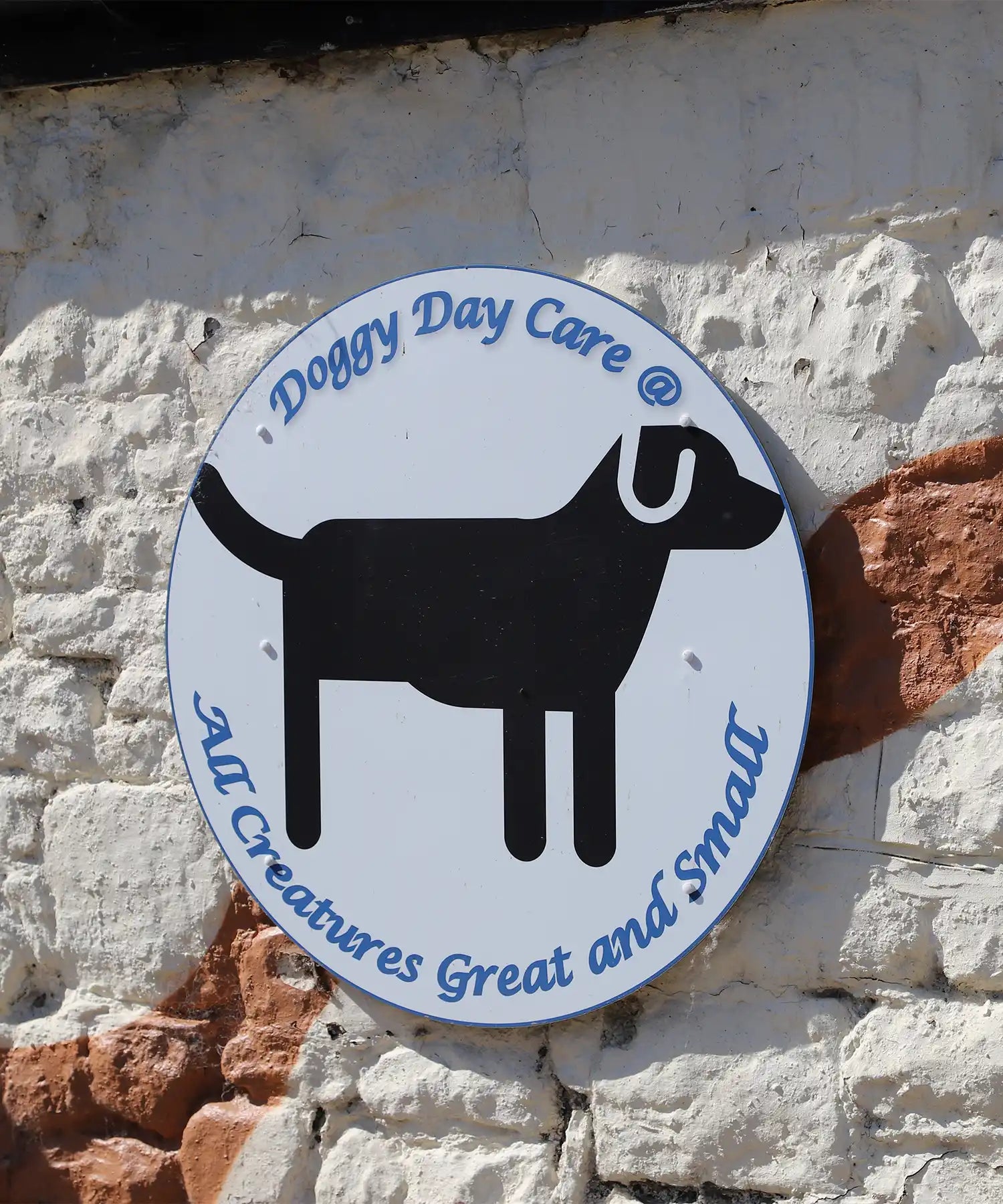 Round sign for “Doggy Day Care @ All Creatures Great and Small” mounted on a textured white and red brick wall, featuring a black dog silhouette.

