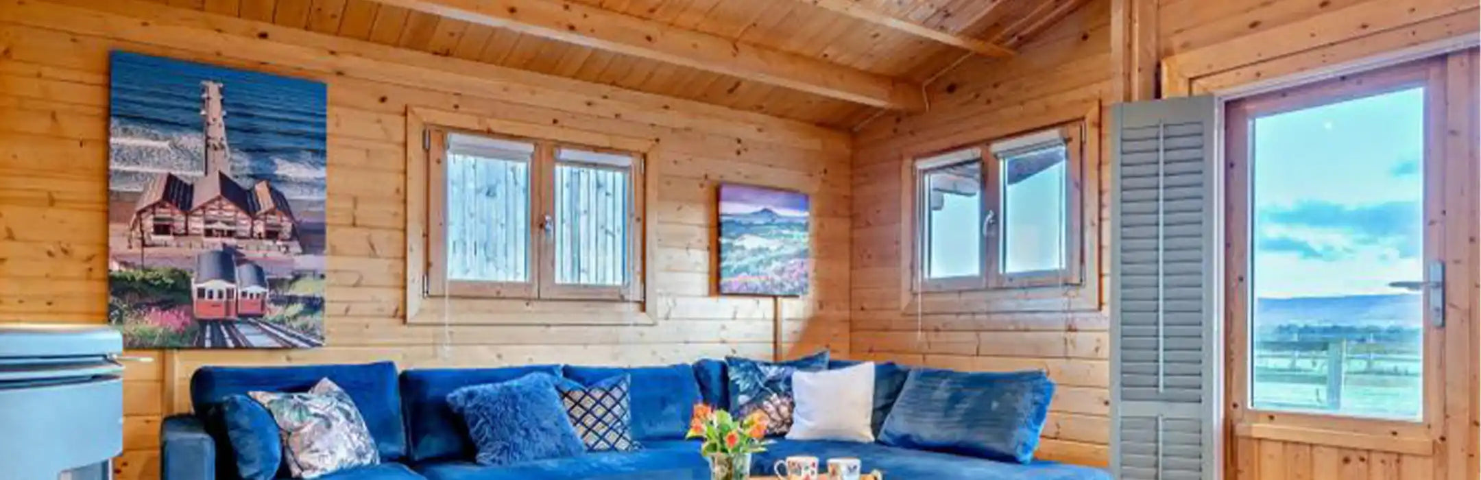 Custom infrared canvas heater panel installed inside a timber lodge living room, featuring blue velvet corner sofa and coastal artwork.